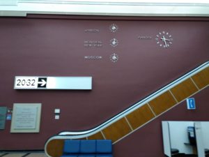 A brown escalator along a bugundy wall featuring four clocks showing the time in London, Montreal and New York, Moscow, and Gander.