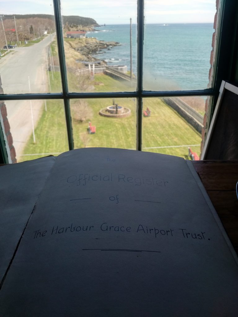The first page of a book, The Official Register of The Harbour Grace Airport Trust in front of a window that overlooks the garden next to the Conception Bay Museum and the ocean.
