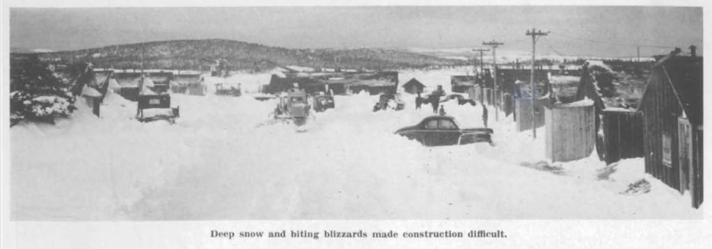 A snowy scene where cars and trucks have snow up over their tires. Buildings are on the right of the picture, and have snow built up around the doors. In the middle of the picture, snowclearing equipment are working.