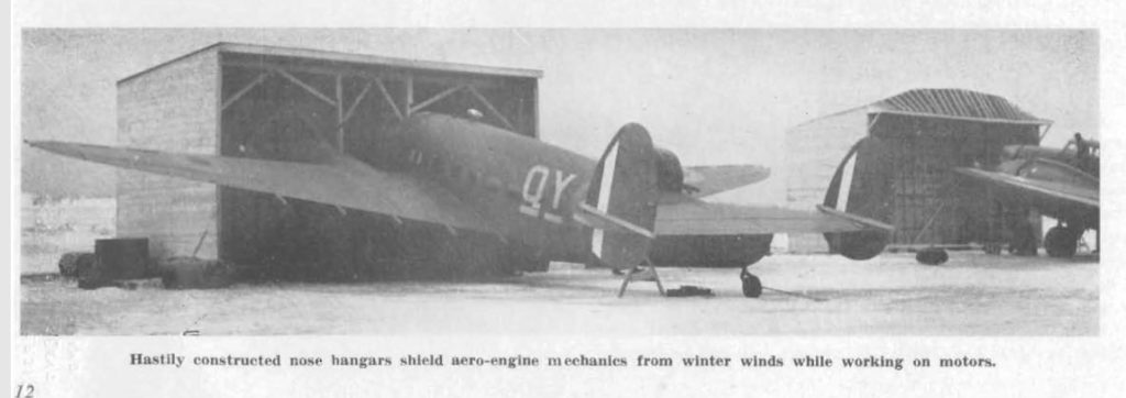 A aircraft is back on to the photo with its nose in a small hangar. The hangar just covers the nose, leaving the wings outside of the structure.