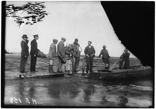 A black and white photo of a small boat tied up at a wharf low to the water on a calm pond. Charles and Ann Lindbergh are standing on the wharf looking toward the boat while a number of men in outfits ranging from suits to military uniforms to work clothes are standing around them and are in the boat. The top left corner of the photo seems to have some scratches and the top right is a black smudge.