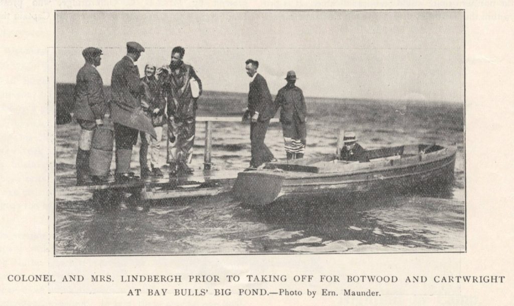 A black and white photo of a small boat tied up next to a wharf that is low to the water of a calm pond. A number of men are on the wharf, and one still in the boat, and in the centre of the photo, on the wharf, are Charles and Ann Lindberg. The caption reads Colonel and Mrs Lindbergh prior to taking off for Botwood and Cartwright at Bay Bulls Big Pond. Photo by Ern Maunder