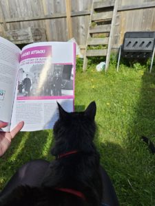 A hand holds open a magazine to an article with the title U-Boat Attack! A black cat is sitting on the person's lap, back of her head visible against the page, as if she is looking at the article. In the background, a green garden and sunshine showing a compost bin and planters, some with strawberry plants visible.