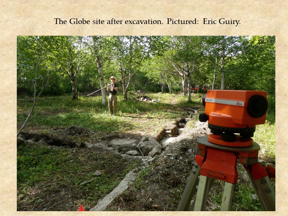 An image of a surveyor's level pointing toward one of the excavation crew who is holding the measuring rod. The area is cleared of trees and bushes