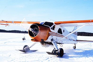 A Gander Aviation plane on skis on the snow. The plane is white with orange markings and the propeller is turning.