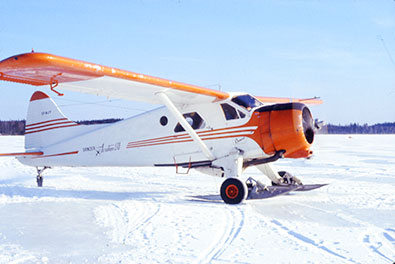 A Gander Aviation plane on skis on the snow. The plane is white with orange markings and the propeller is turning.