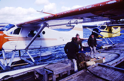Three men on the end of a dock next to a white float plane with orange markings. They are standing under the wing. A yellow float plane is in the background.
