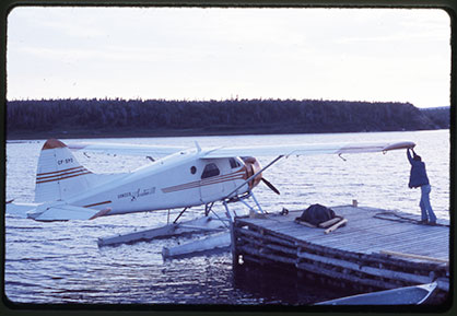 A white float plane with orange markings is on a pond at the end of a dock. A man is reaching up to the wing that is over the dock.