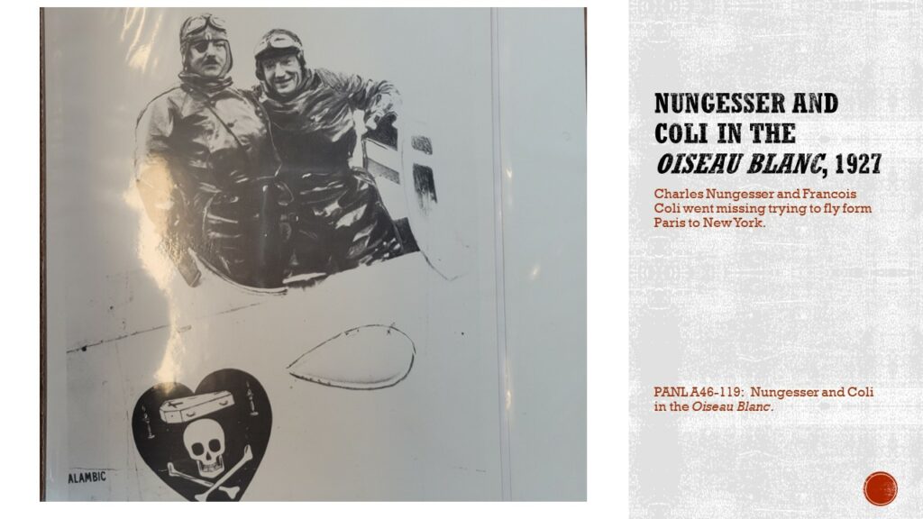 Black and white image of two aviators in winter flying suits in a white aircraft. On the side of the aircraft is a black heart with a coffin and skull and cross bones.

Text: Nungesser and Coli in the Oiseau Blanc, 1927. Charles Nungesser and Francois Coli went missing trying to fly from Paris to New York.

Caption: PANL A46-119: Nungesser and Coli in the Oiseau Blanc.