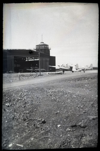 A black and white image of a building under construction and two aircraft near the building. Most of the image is of the ground and a path leading to the aircraft, putting the building and aircraft further into the background so details are unclear.
