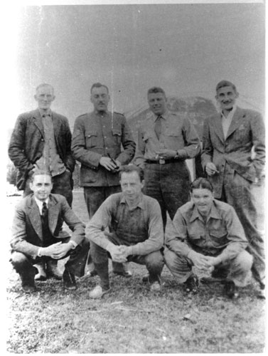 A black and white photo of seven men. Four are standing behind three who are crouched before them. They are in various outfits, from Newfoundland Ranger and RAF uniforms to suits, to casual clothes.
