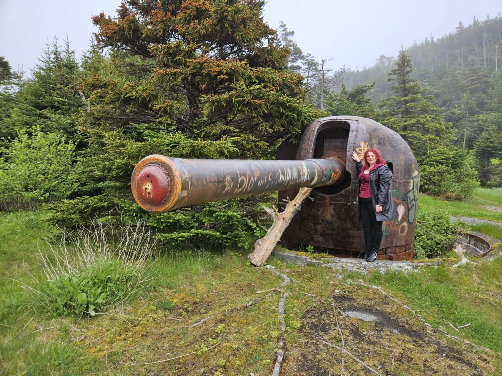 A colour image of the author standing next to a large gun. The gun is rusted and covered in graffiti, and the end is blocked. The gun is significantly larger than the author, and the whole area is surrounded by trees.