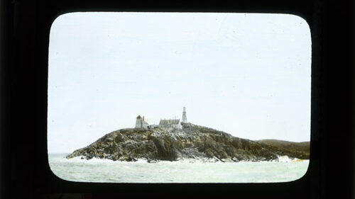 A lightly coloured photo of a lighthouse and outbuildings on a rugged, rocky, coast surrounded by ocean.