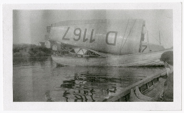 A black and white image of the wing of the Bremen and other parts of the aircraft on a boat. The letters and numbers on the wing are upside down, but read D 1167. The serial number is reflected in the water. There is a person in the foreground looking at the aircraft pieces, and land with buildings in the background.