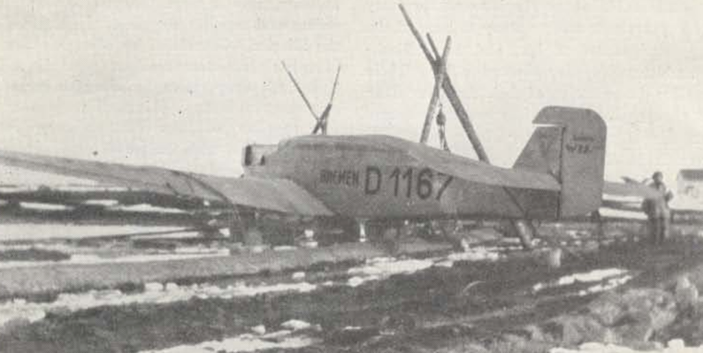 A black and white image of an aircraft from an angle behind the plane that shows the tail, fuselage, and one wing. The wing looks like it is being suspended by pulleys on two different systems of tall logs wrapped to form pyramid structures. There is an unidentified man on the wing in the background of the image, and a house far in the distance. The marking on the fuselage are visible and read Bremen D1167"