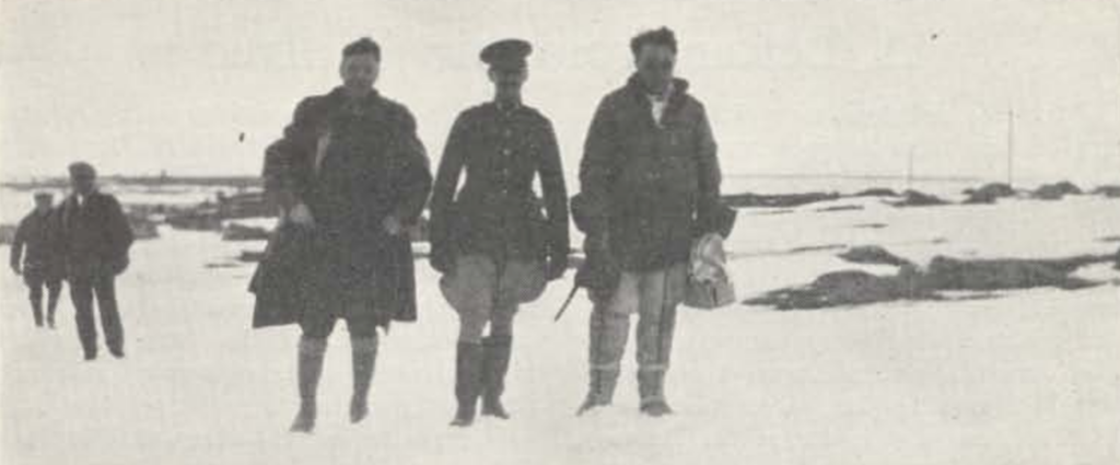 Three men stand in the centre of the black and white photo. They seem to be in motion, walking towards the camera. Further back and to the left are two more people, also moving toward the camera. The land is barren and snow-covered with some rocks visible through the snow.