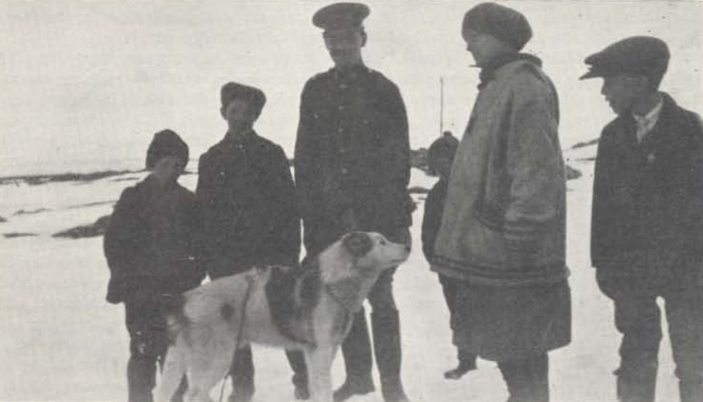 A black and white photo of 6 people and a dog. Three of the people on the left, two children and a man, are looking at the camera. A woman and another child are to the right and they are looking at something to the left, out of the shot. There is a small child behind the woman, leaning to look past her at the camera. The dog is standing in front of the three looking at the camera, looking to the right, towards the woman.