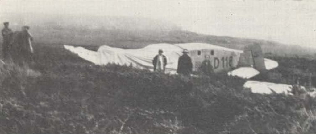 A black and white photo of the Bremen on land that seems mostly composed of short shrubs. The nose of the aircraft is covered by some sort of sheet. There are unidentifiable people both in front of the aircraft and to the left.