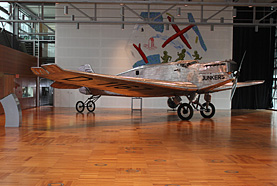 A colour photo of the small aircraft. The aircraft is mostly silver in colour and "Junkers" is written along the nose. It is in an exhibit setting.